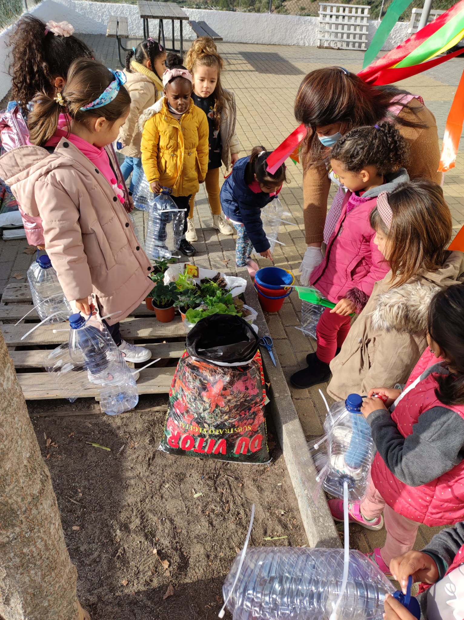 A foto mostra a preparação e distribuição do substrato em garrafões de água de plástico, para hortícolas e aromáticas. Trabalho com alunos do Jardim de Infância.