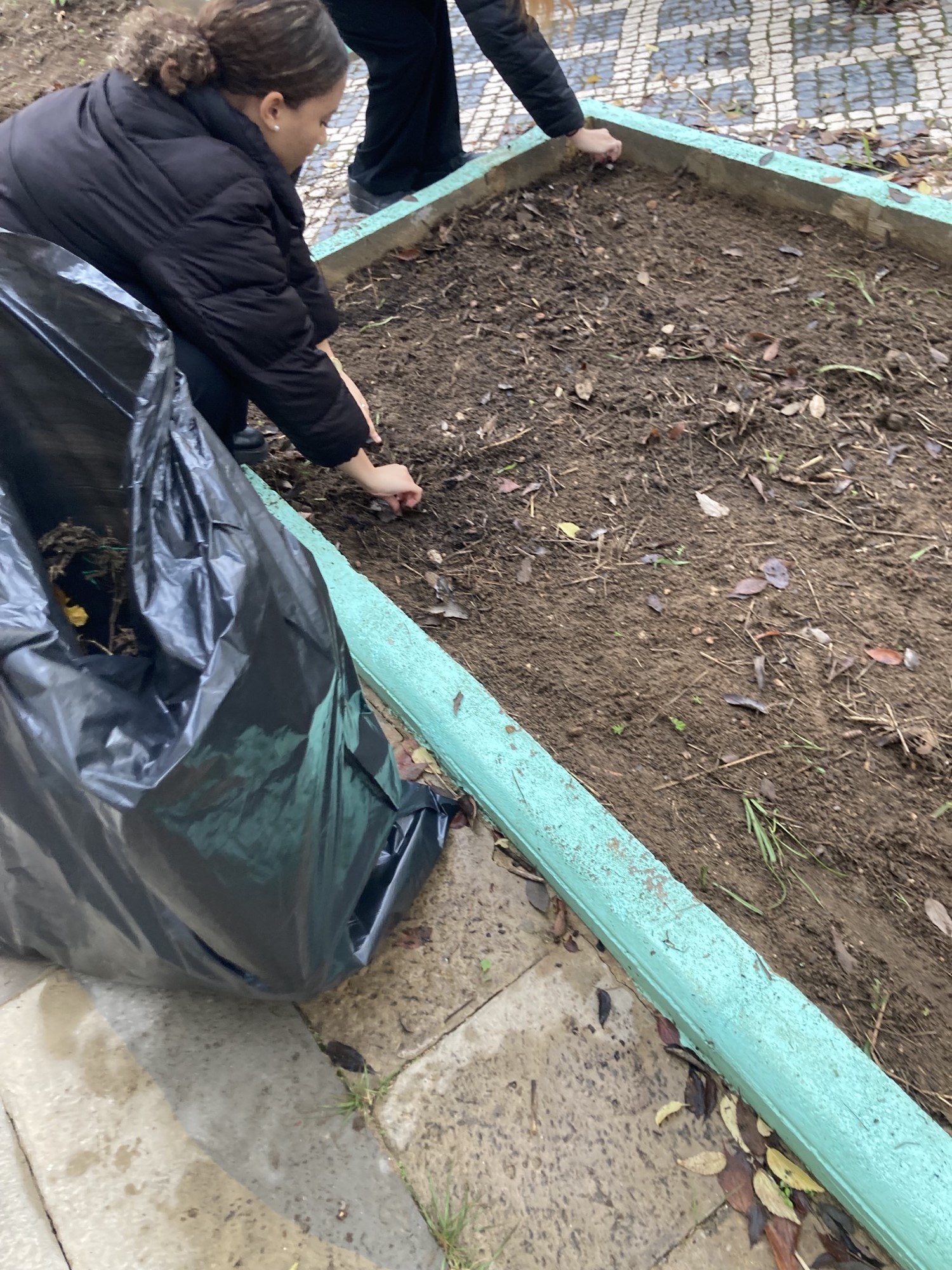 A aluna está a realizar a limpeza da horta com grande cuidado e atenção aos detalhes. Ela dedica-se a retirar as partículas mais pequenas, como plásticos e beatas, para garantir que o solo esteja em perfeitas condições para as futuras plantações. Este tra