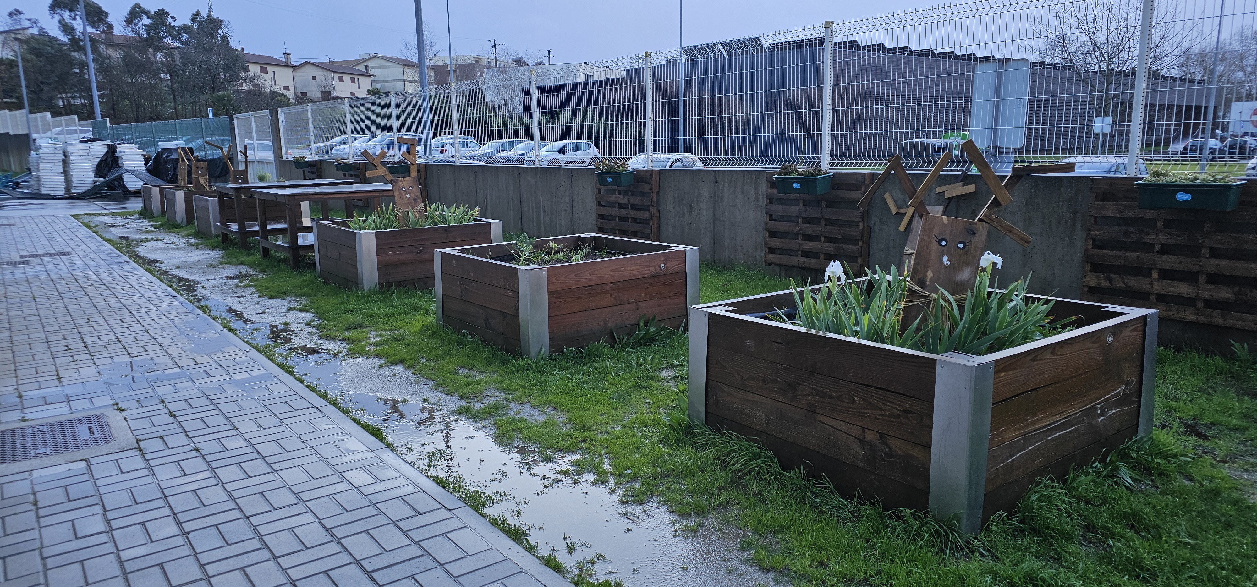 Nesta fotografia temos a imagem das 6 caixas de plantio e da forma como estão dispostas no espaço. Também são visíveis 2 bancadas em madeira, que servem de suporte às diversas atividades a realizar.