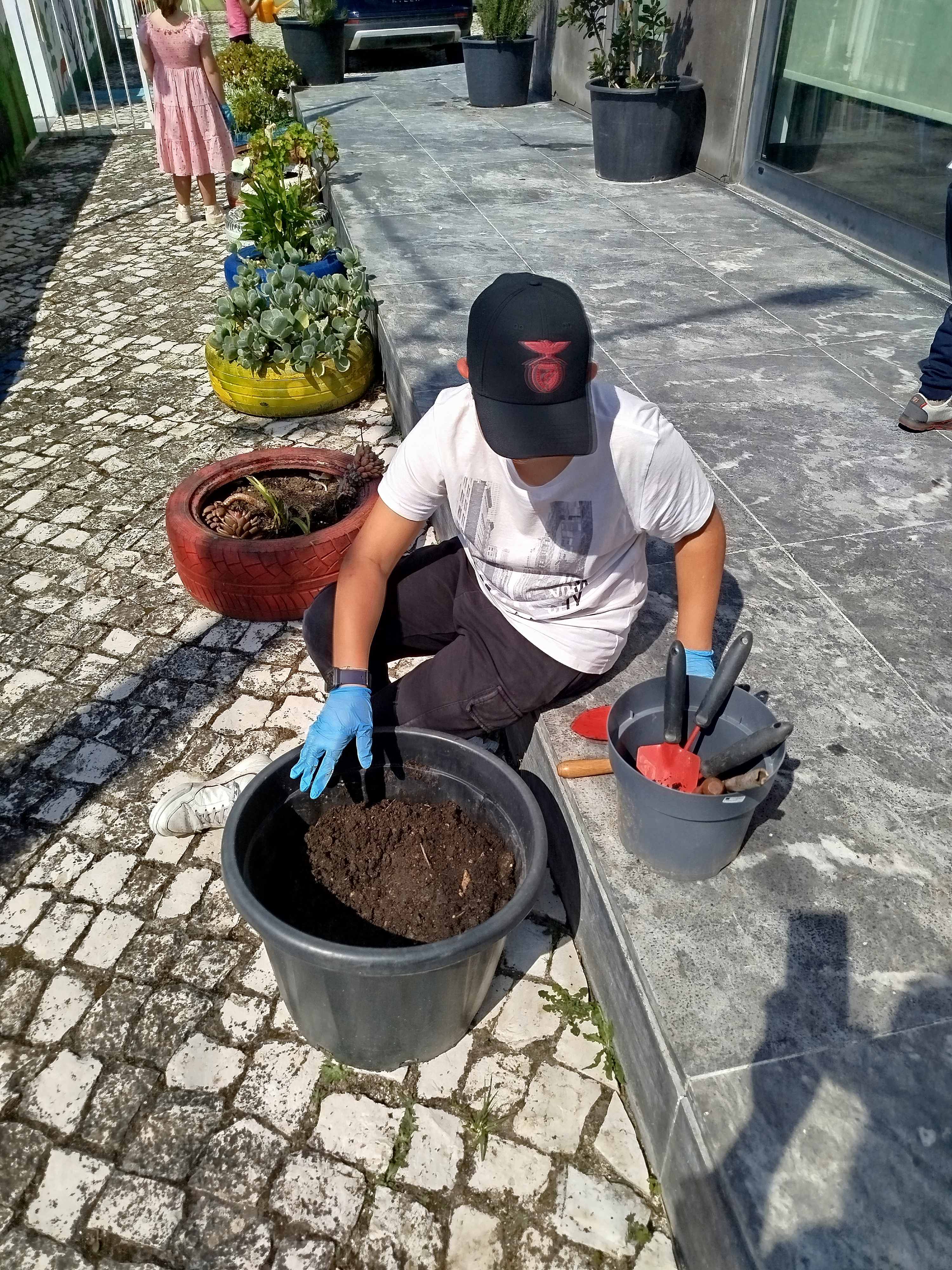 Criança a manusear a terra num balde, preparando o substrato para posterior plantação de plantas aromáticas na horta escolar.