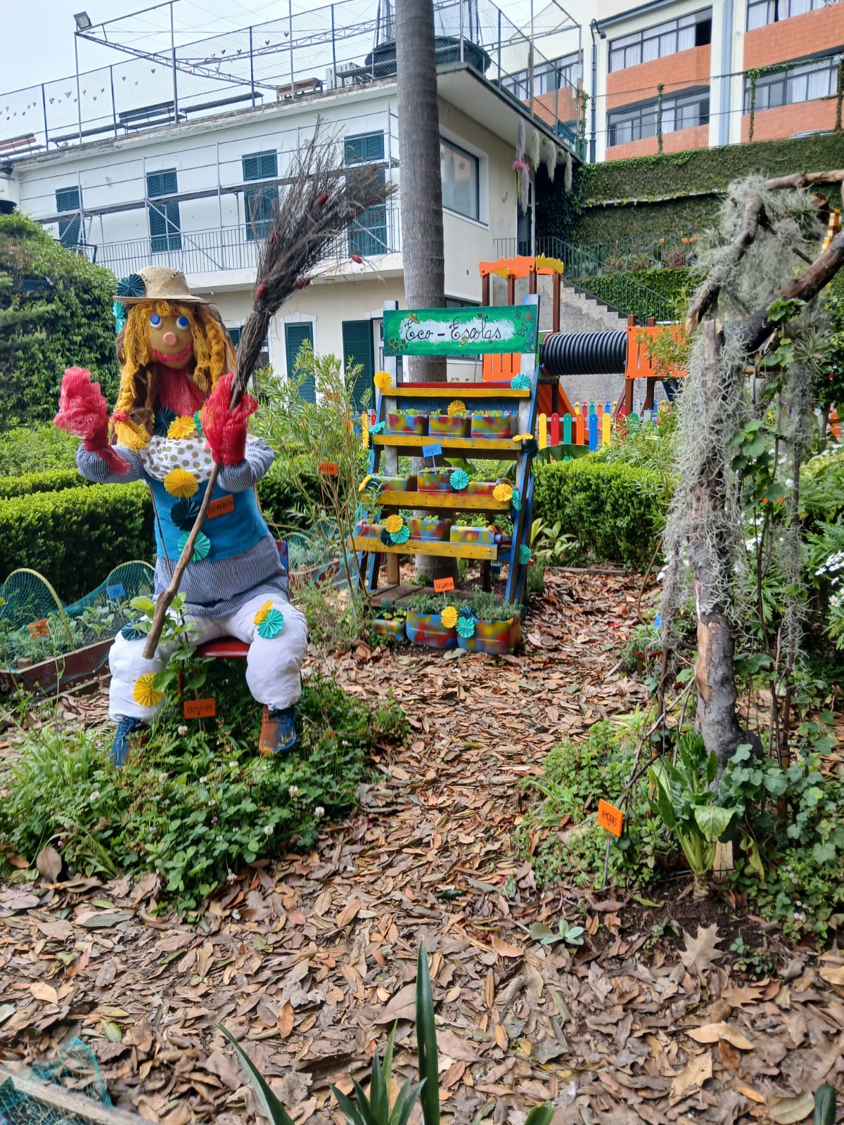 A fotografia mostra a lateral da nossa horta. É possível observar o espantalho construído pelos alunos e a estrutura de madeira onde foram plantadas alfaces e ervas aromáticas.